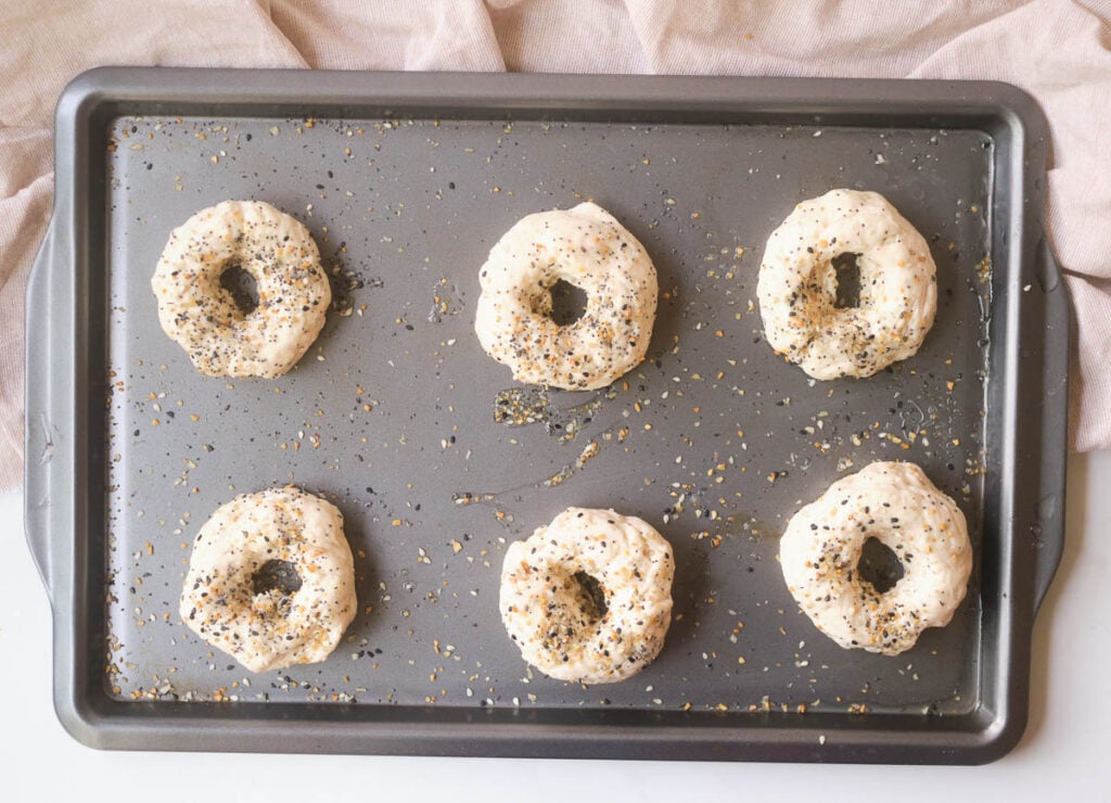 Pre-baked bagels on baking sheet topped with seasoning.