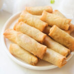 Cooked air fryer frozen spring rolls stacked on white plate.