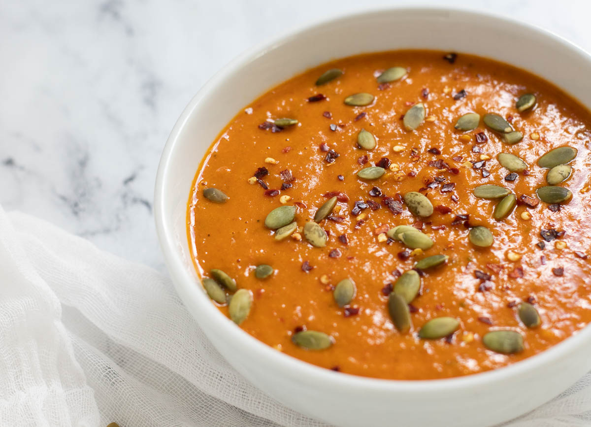 Roasted butternut squash and red pepper soup in white bowl, topped with red pepper flakes and pumpkin seeds.