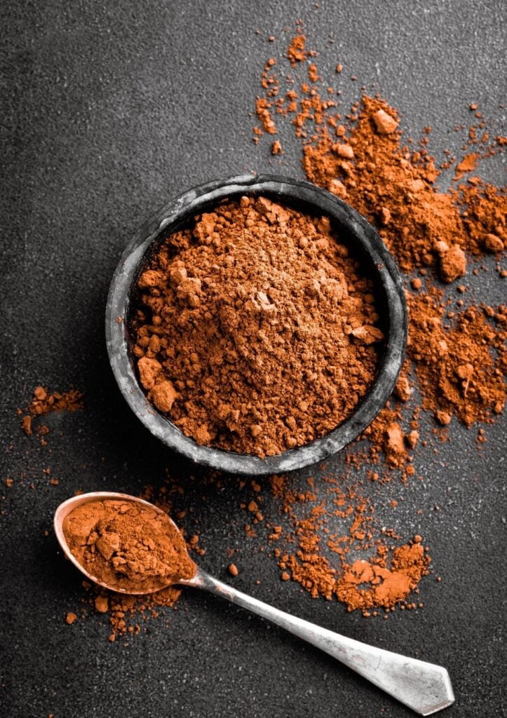 Cocoa powder in small gray bowl beside spoon with cocoa powder.