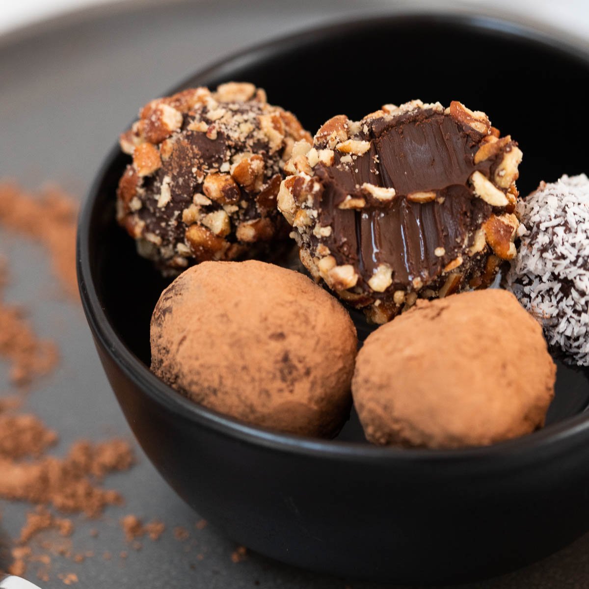 Chocolate vegan truffles coated in nuts, cacao powder, and coconut in small black bowl.