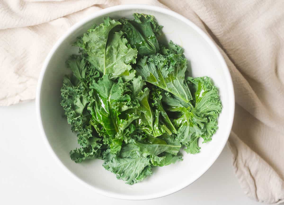 Kale leaves topped with olive oil in white mixing bowl. 