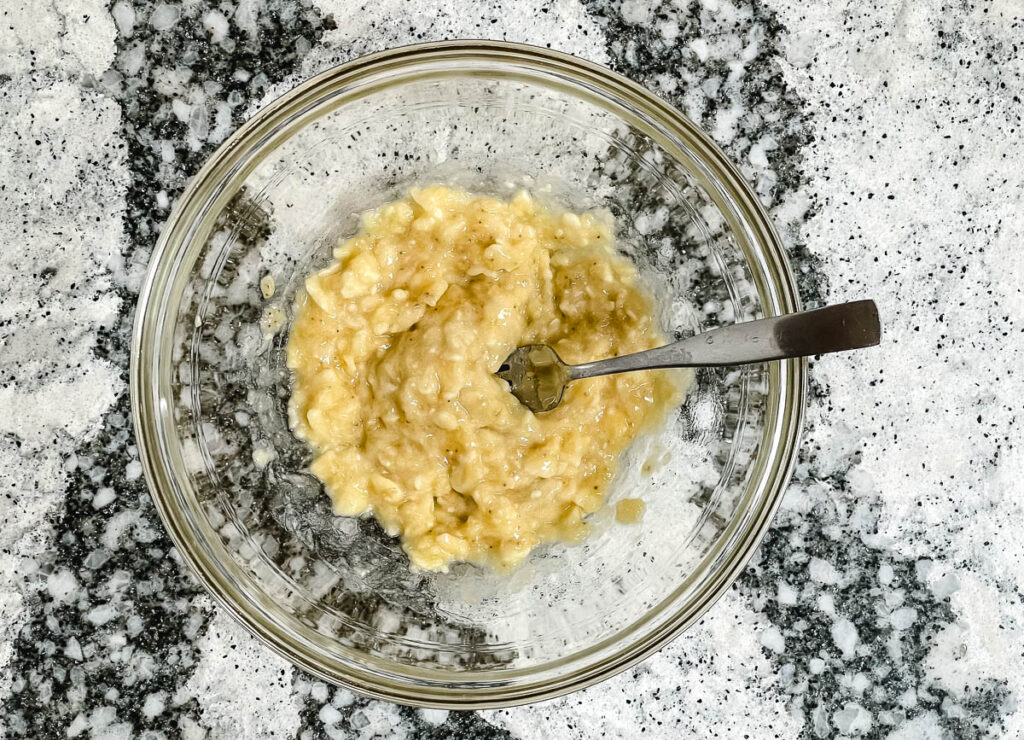 Mashed banana with fork in mixing bowl. 