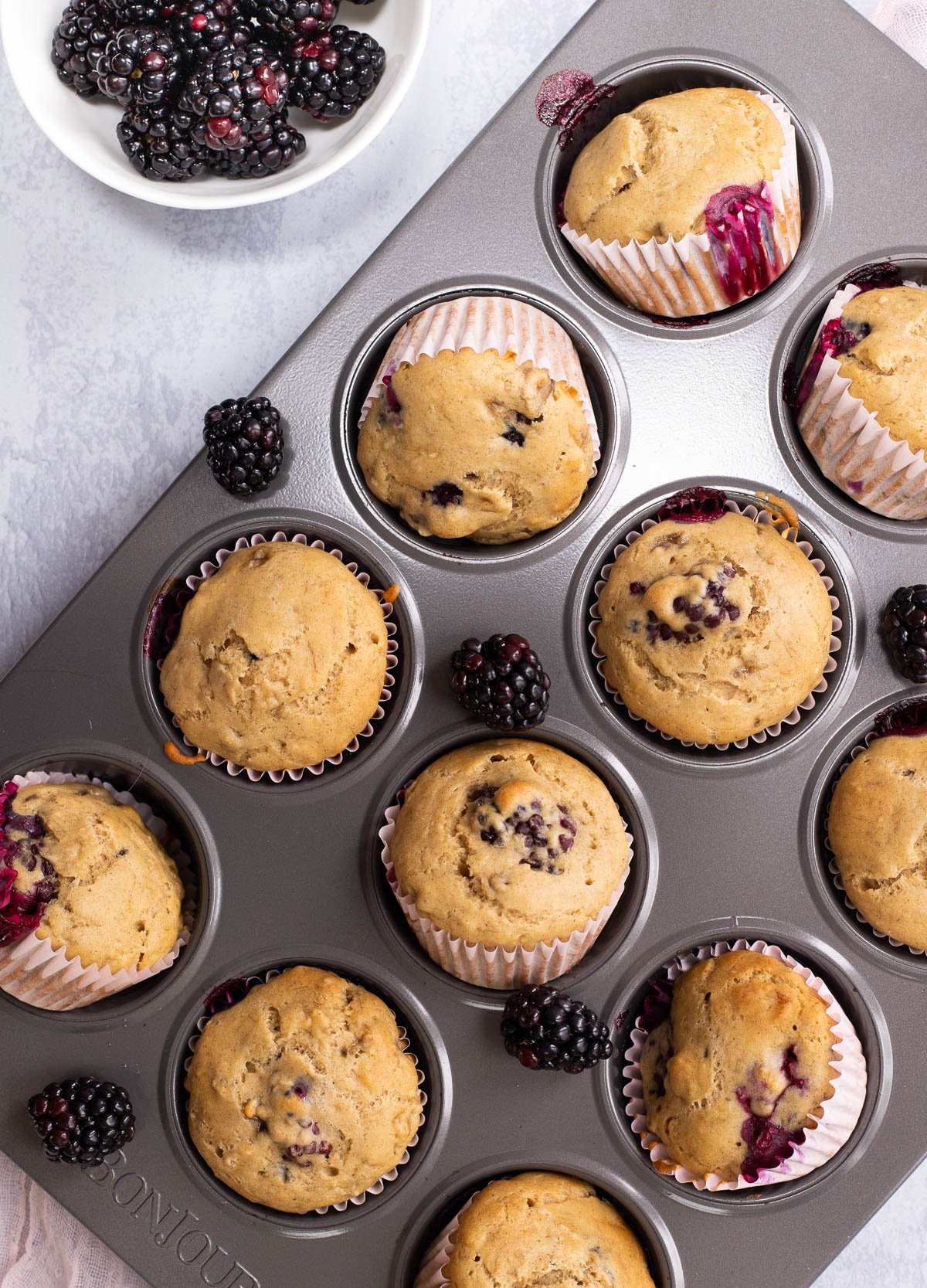 Vegan blackberry muffins in muffin tin surrounded by fresh blackberries.