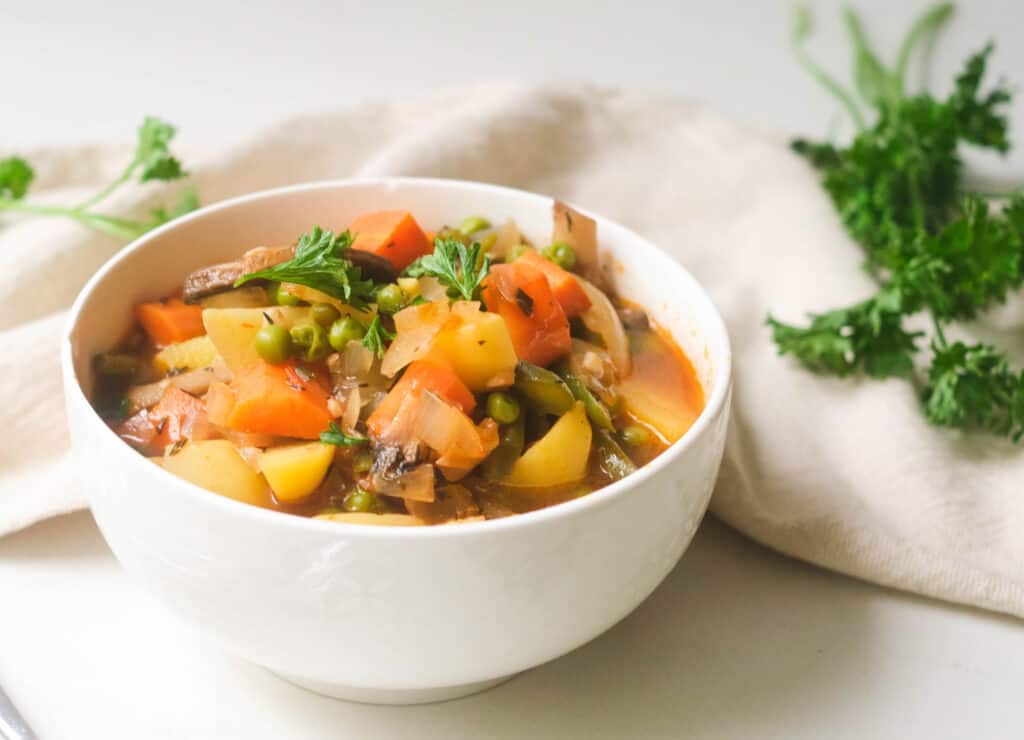 Bow of vegetarian vegetable stew with carrots and potatoes in white bowl, topped with parsley.