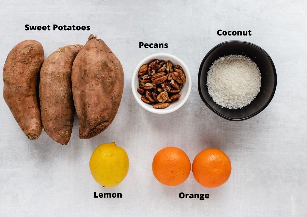 Three sweet potatoes, bowl of coconut flakes, bowl of pecans, lemon, and two small oranges.
