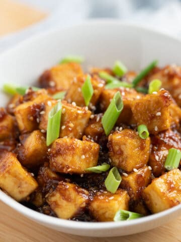 Bowl of pan-fried tofu cubes coated in teriyaki sauce and topped with scallions.