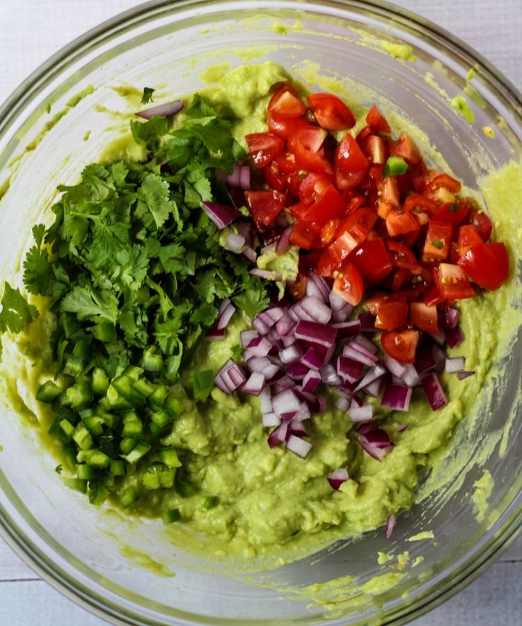 Bowl with mashed avocado, diced red onion, diced cherry tomatoes, chopped cilantro, and diced jalapeño peppers.