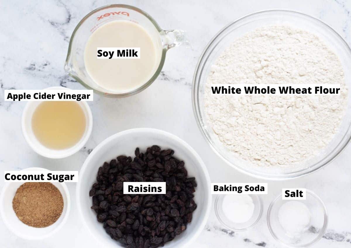soy milk in measuring cup, flour in glass bowl, raisins in white bowl, coconut sugar and apple cider vinegar in small white bowls, and salt and baking soda in small glass bowls