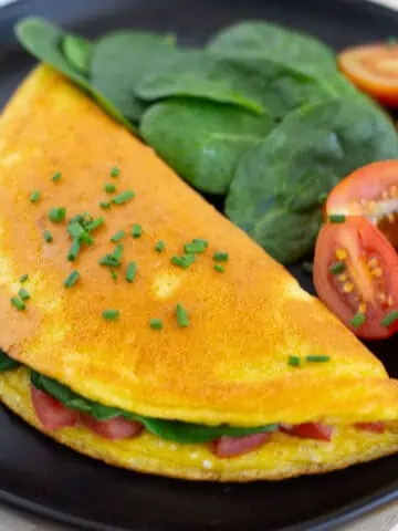 Just Egg omelette on black plate served with a side of spinach and sliced tomatoes.