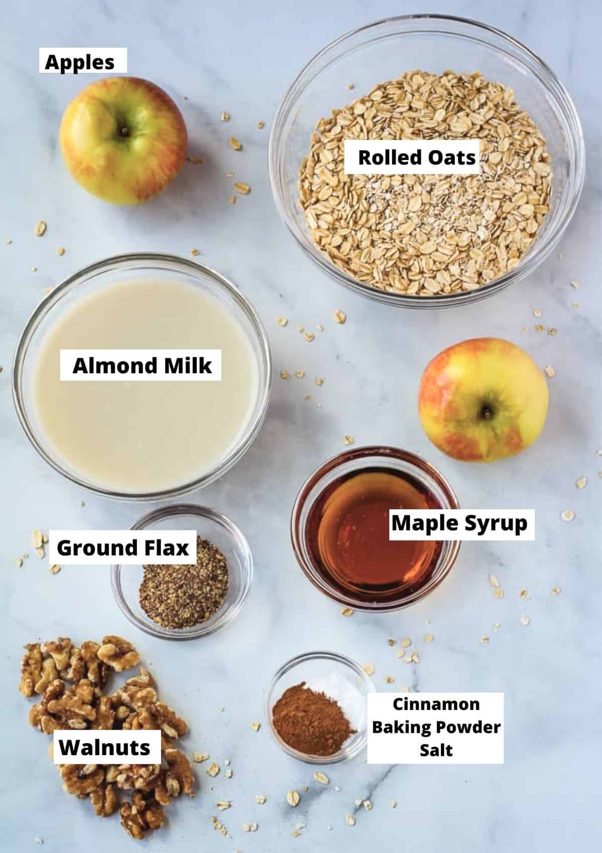 Ingredients for baked oatmeal including rolled oats, apples, almond milk, maple syrup, walnuts, ground flax, cinnamon, baking powder, and salt. 