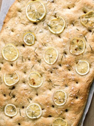 Loaf of baked vegan focaccia with slices of lemon and fresh rosemary on cutting board.
