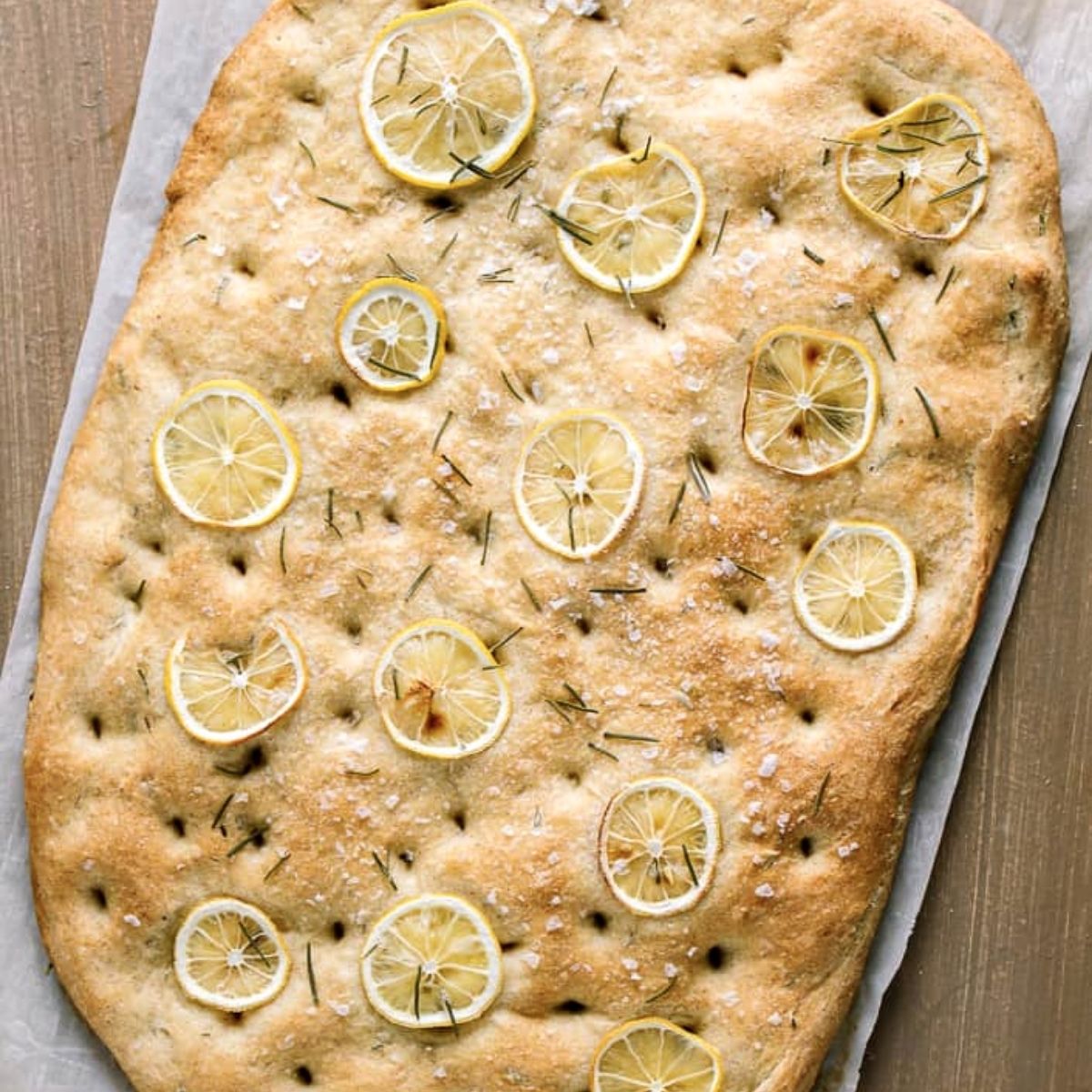 Loaf of baked vegan focaccia with slices of lemon and fresh rosemary on cutting board. 