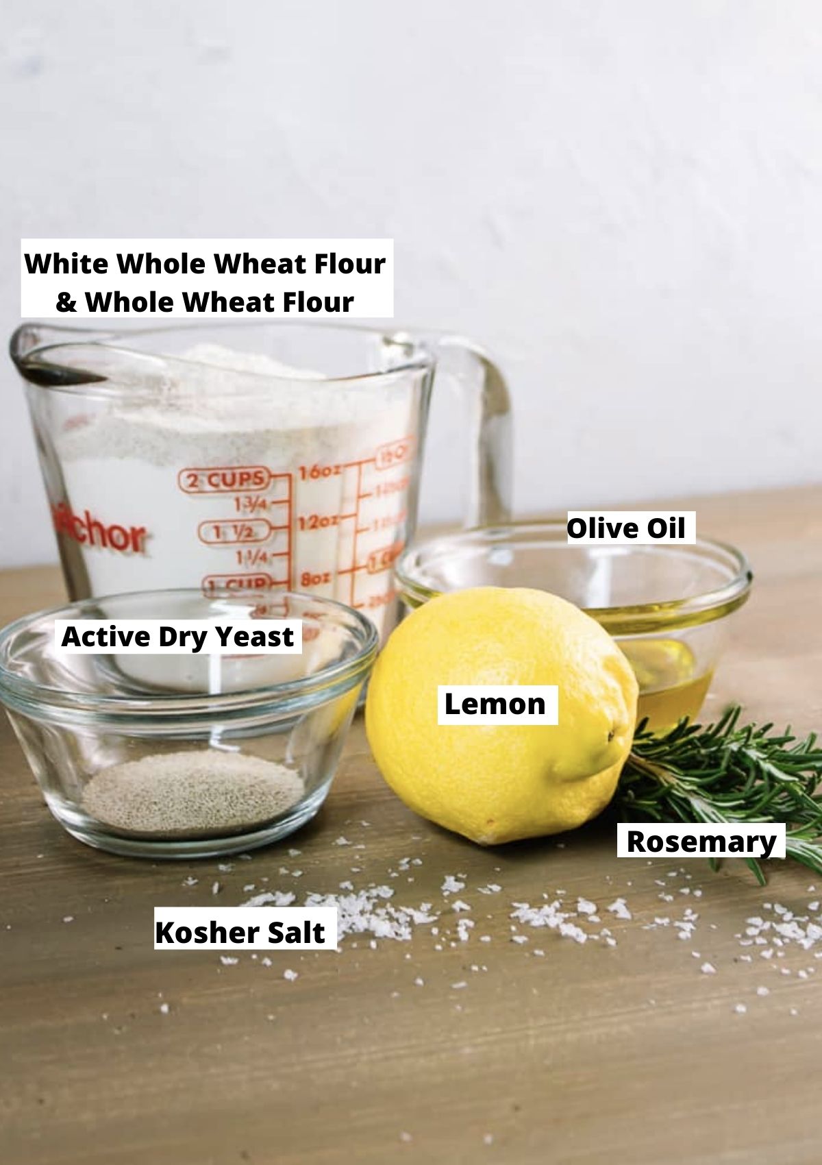 Cup of flour, bowl of dry yeast, olive oil in a bowl, lemon, fresh rosemary, and salt on table top. 