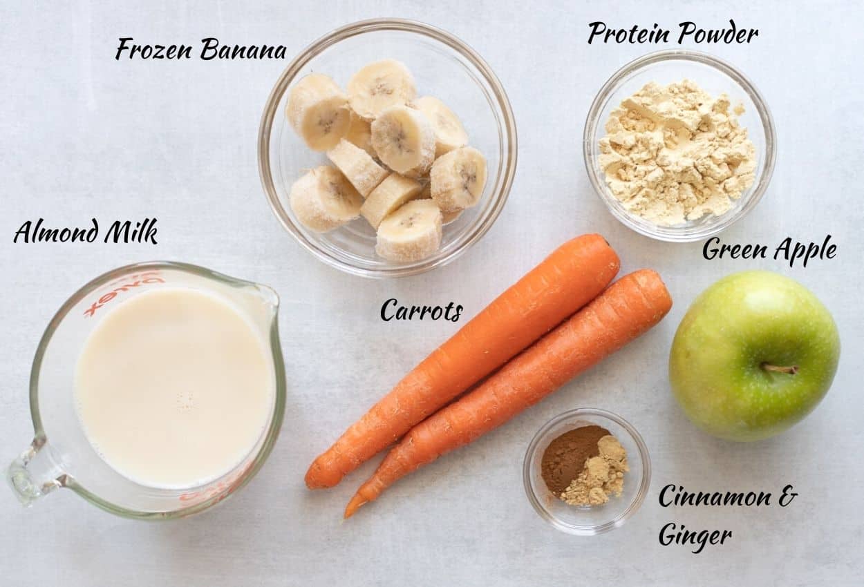 Frozen bananas in glass bowl, protein powder in glass bowl, green apple, cinnamon and ginger in glass bowl, two carrots, almond milk in glass measuring cup. 
