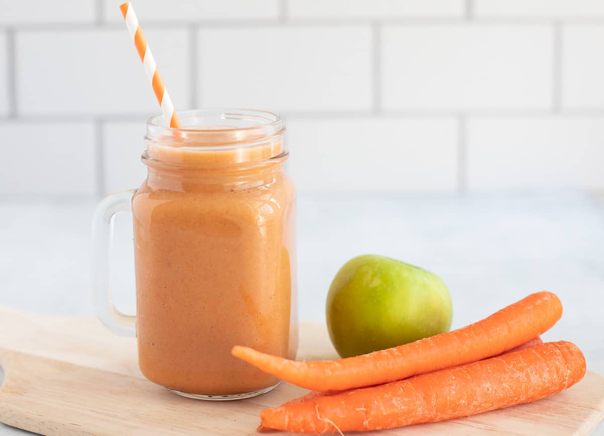 Apple carrot smoothie in glass mug beside a stack of carrots and a green apple.