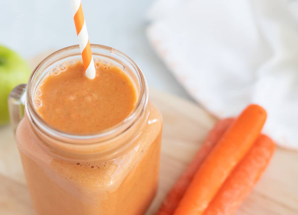 Carrot smoothie in glass mug, beside a stack of carrots. 