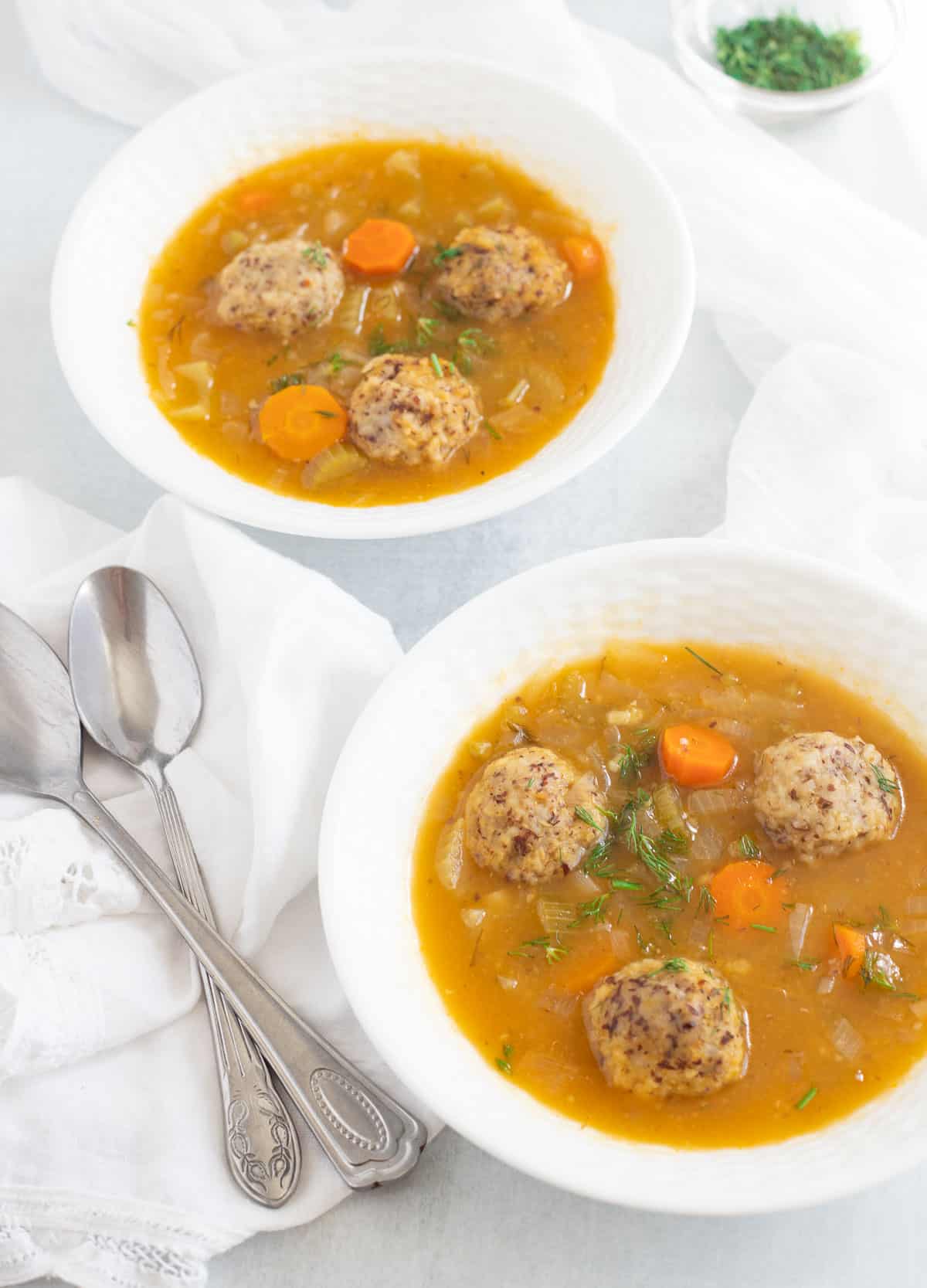 Matzo ball soup in two white bowls, with spoons beside them. 