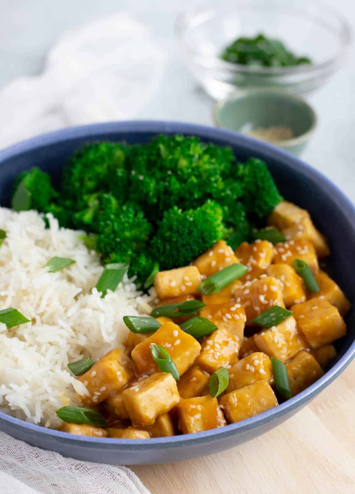 Tofu vegan orange chicken served in a blue bowl with white rice and broccoli. 