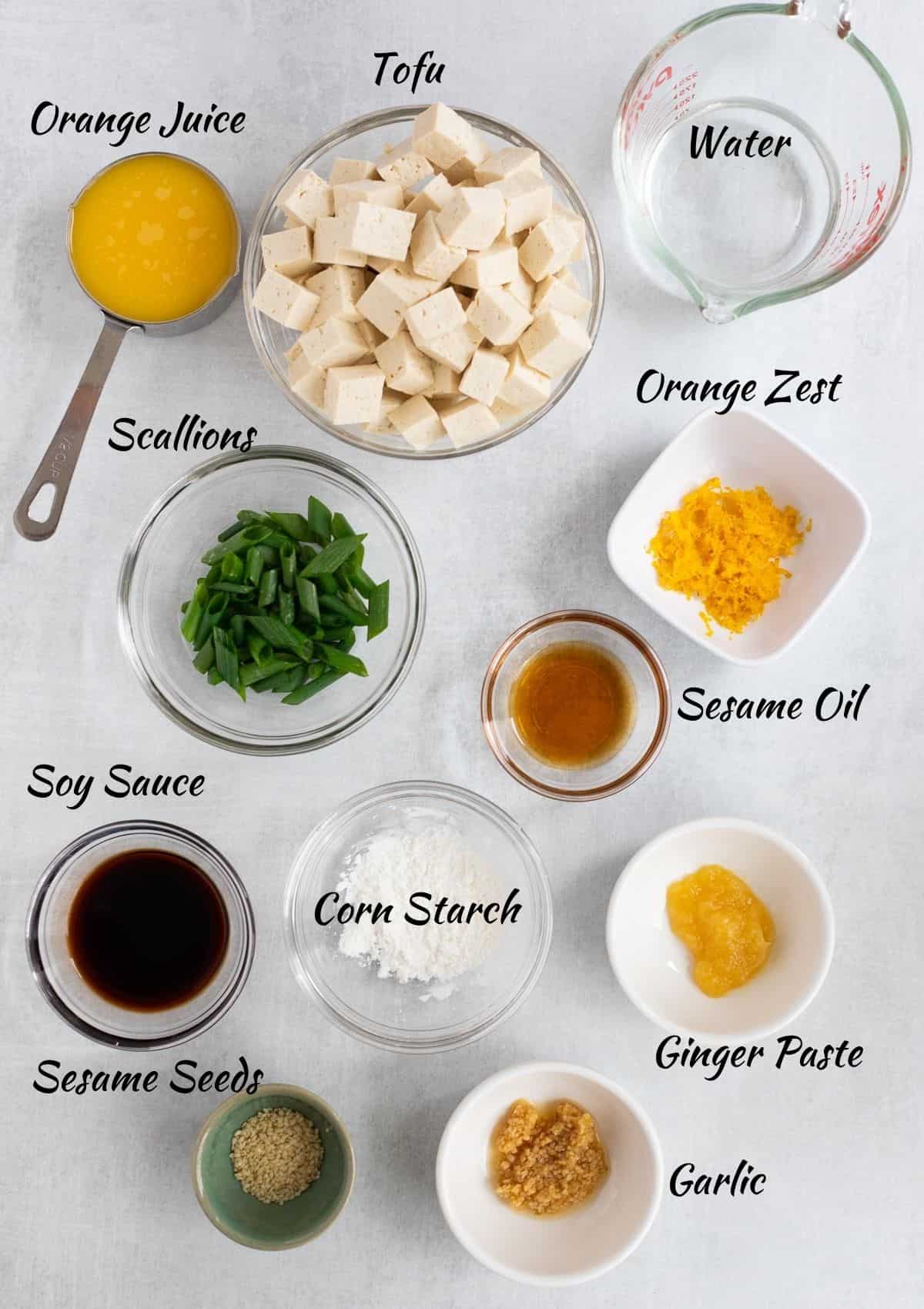 Orange juice in measuring cup, cubed tofu in glass bowl, water in measuring  cup, orange zest in small square bowl, chopped scallions in bowl, sesame oil in bowl, soy sauce in bowl, corn starch in bowl, ginger paste in bowl, sesame seeds in bowl, minced garlic in bowl. 