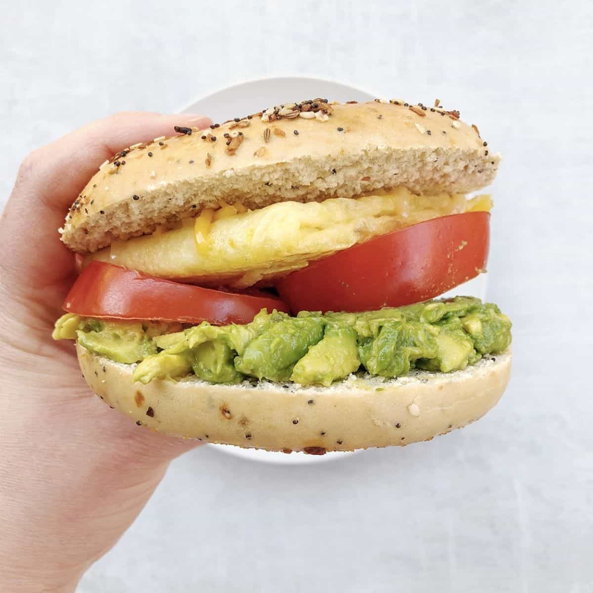 Hand holding vegan bagel sandwich with egg, tomato, and avocado. 