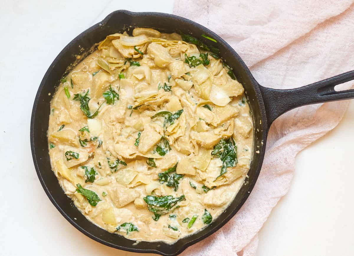 Cashew cream poured over artichokes and spinach in skillet. 
