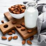 Almond milk in jug on cutting board surrounded by raw almonds.