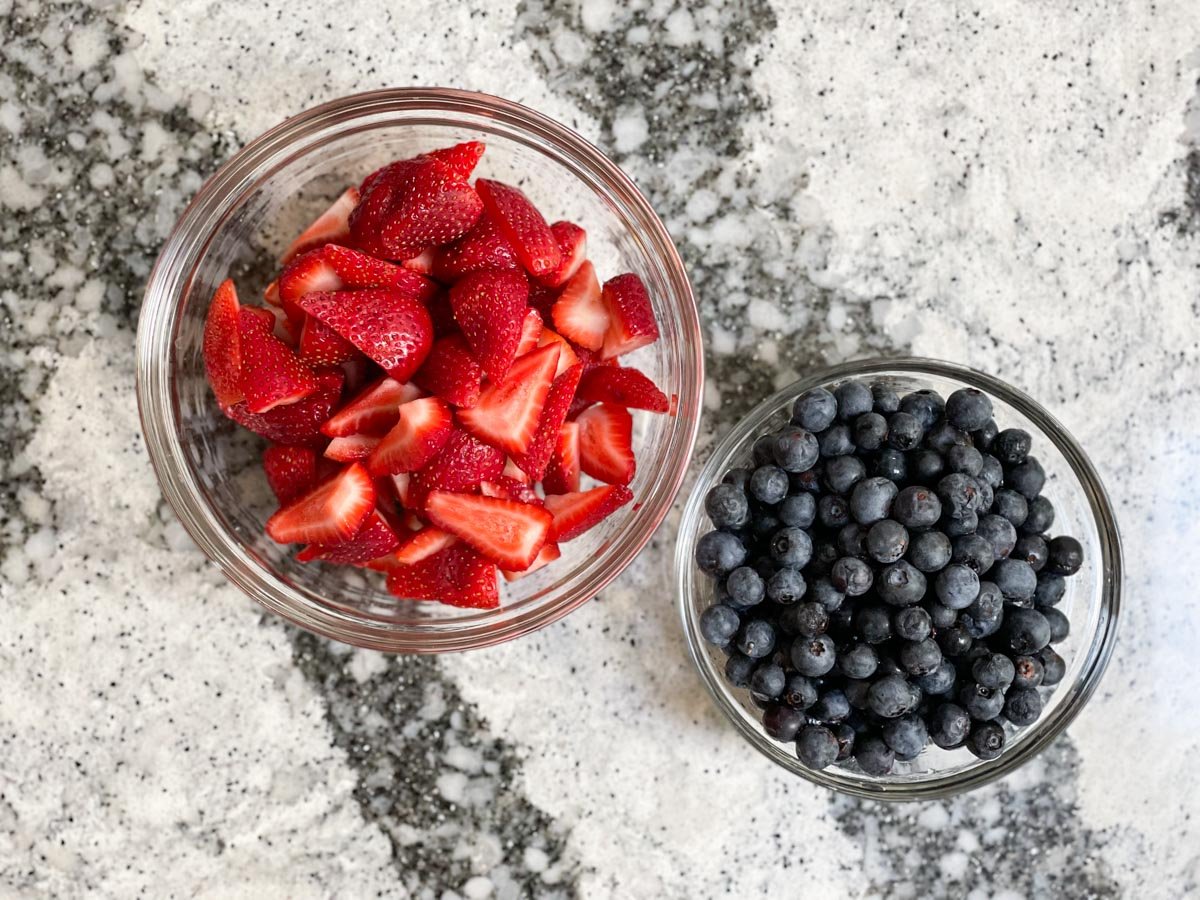 Sliced strawberries in a bowl beside washed blueberries.