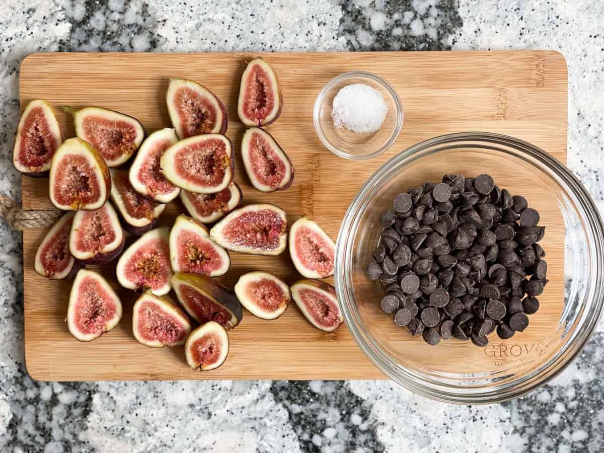 Sliced figs on cutting board beside bowl of chocolate chips, and small bowl of salt. 
