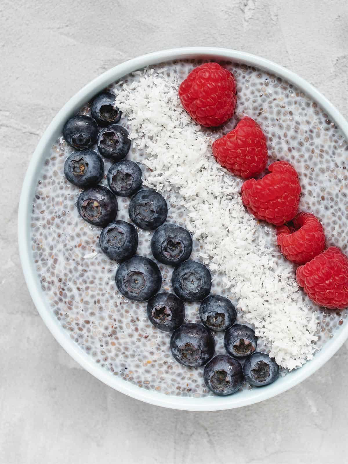 Chia pudding in white bowl topped with blueberries, coconut flakes, and raspberries.
