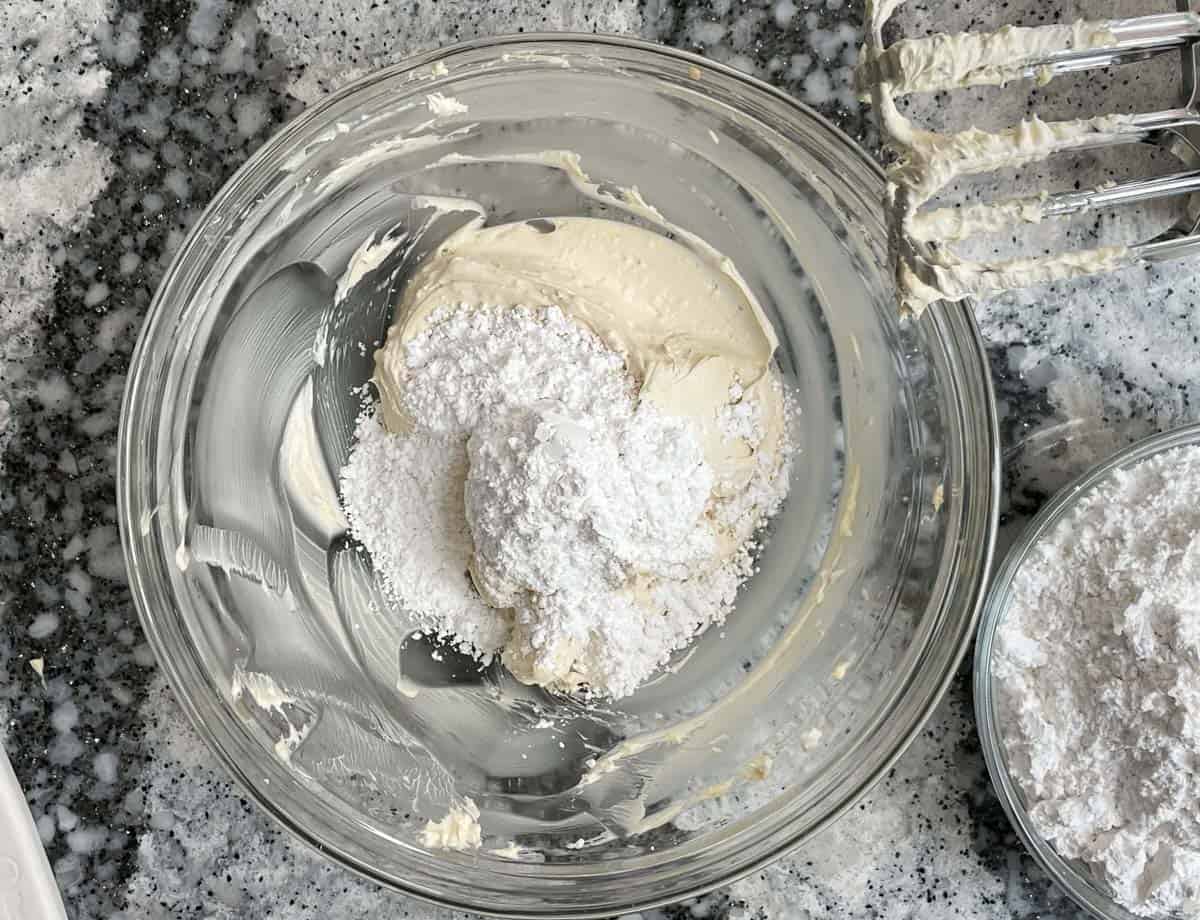 Adding powdered sugar to whipped butter and cream cheese in glass bowl.
