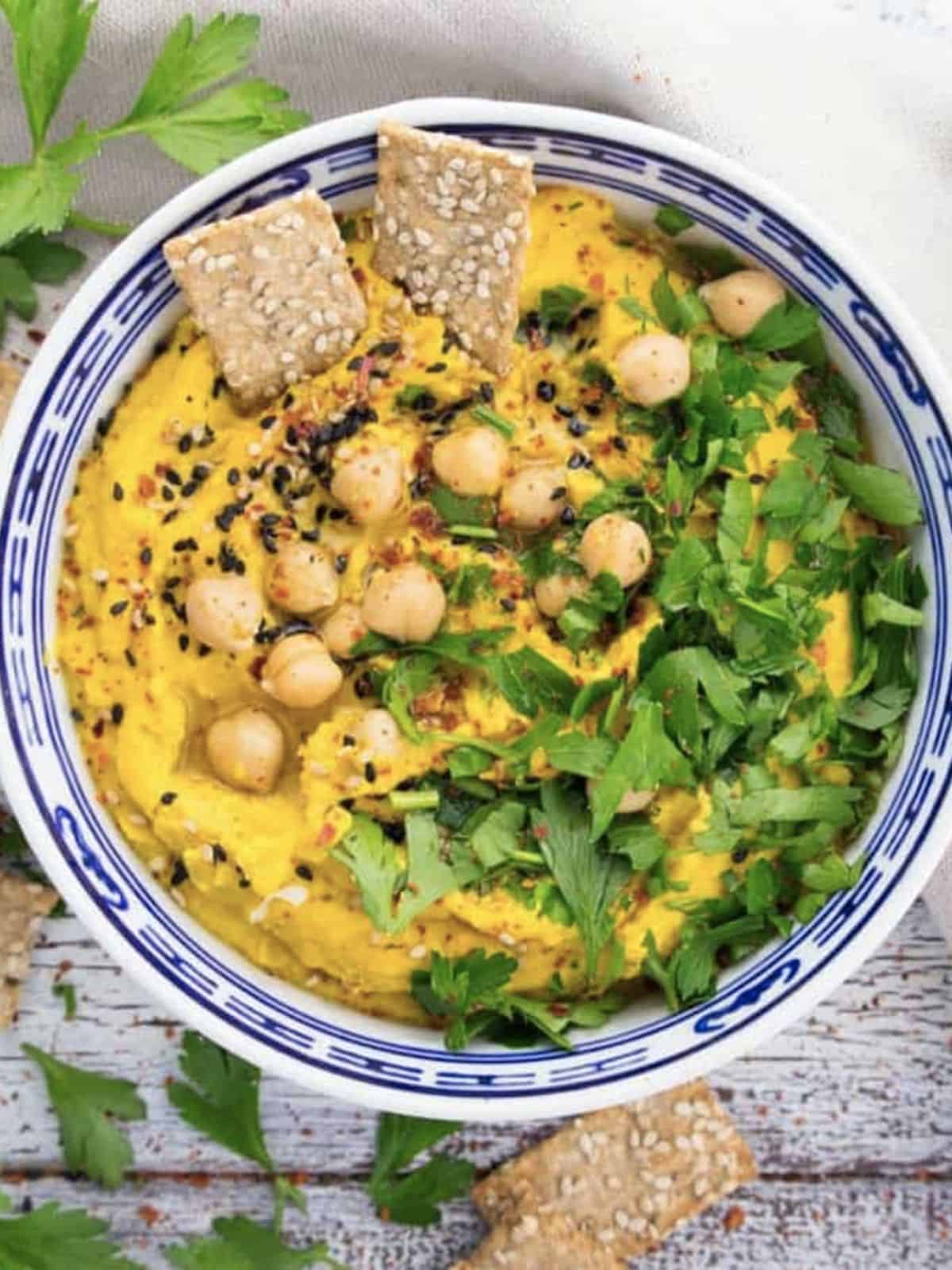Pumpkin hummus in bowl topped with with fresh herbs, chickpeas, and seeds. 
