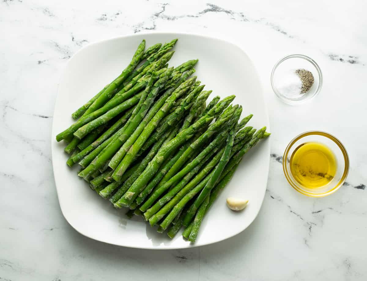 Frozen asparagus spears on plate, olive oil, salt, pepper, and garlic clove.
