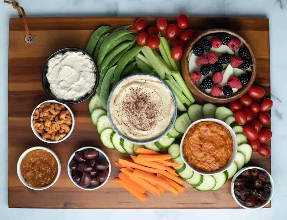 Cucumbers, tomatoes, carrots, celery, snap peas, and small bowls filled with dates, olives, jam, and nuts on wood serving board. 