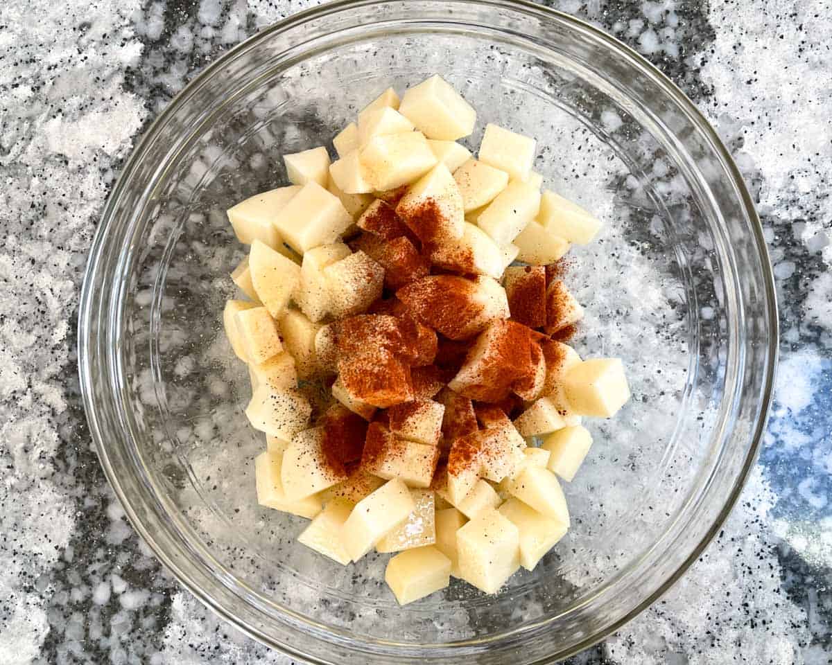 Diced and peeled potatoes topped with spices in glass mixing bowl.
