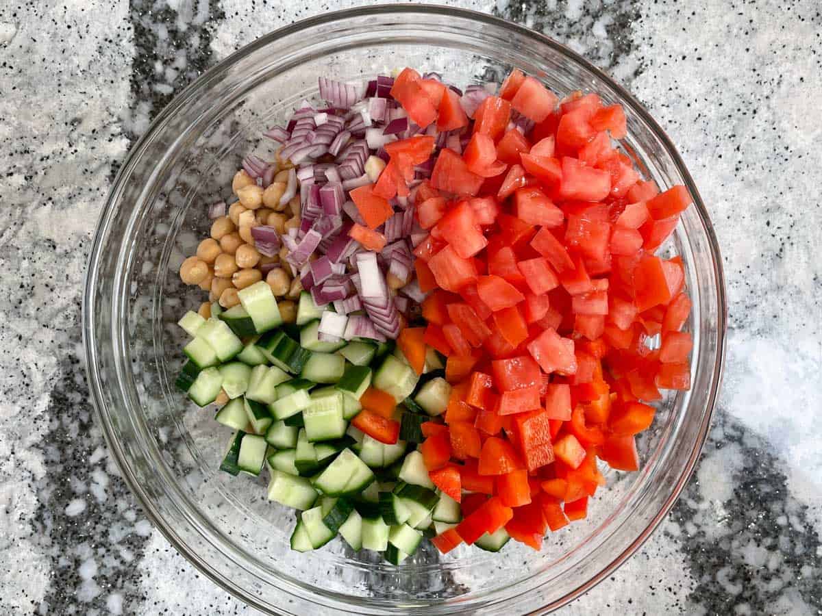Large glass bowl filled with cucumbers, chickpeas, red onion, diced tomatoes, red onions, and quinoa. 