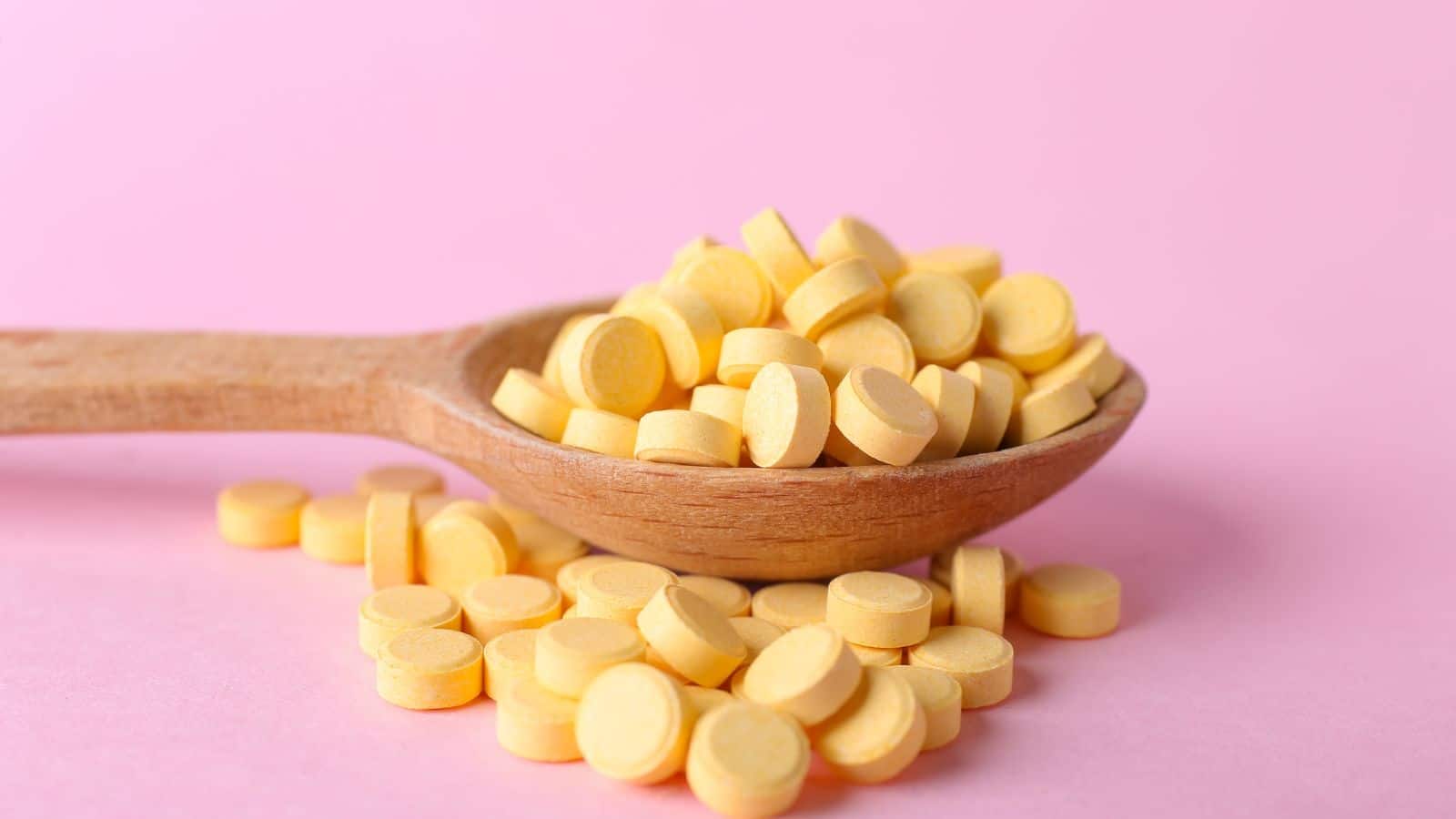 Pile of vitamins on a wooden spoon against a pink background.
