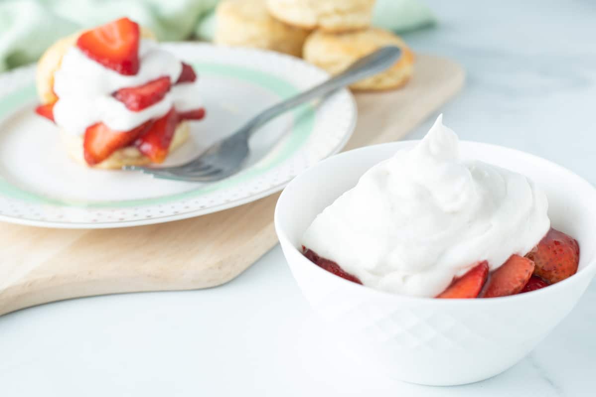 Strawberries in a bowl topped with vegan whipped cream, and strawberry shortcake in the background.
