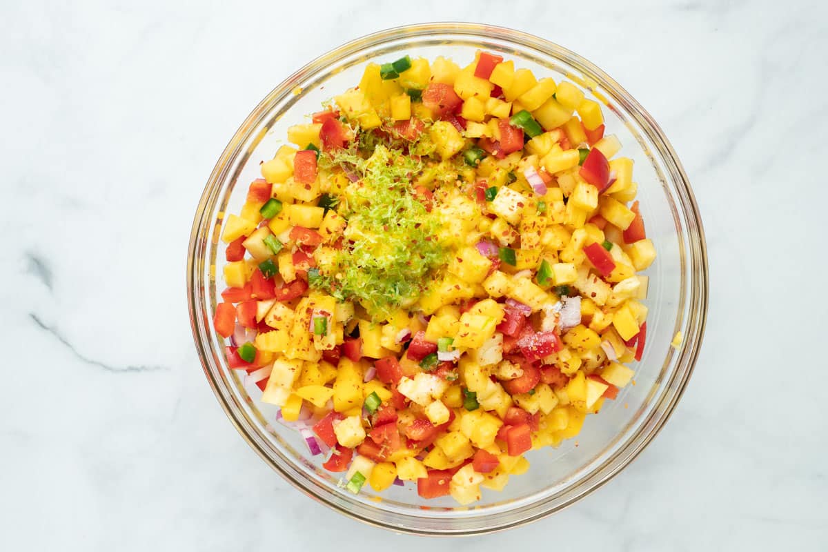 Pineapple mango salsa in a large glass bowl topped with lime zest, salt, and red pepper flakes.
