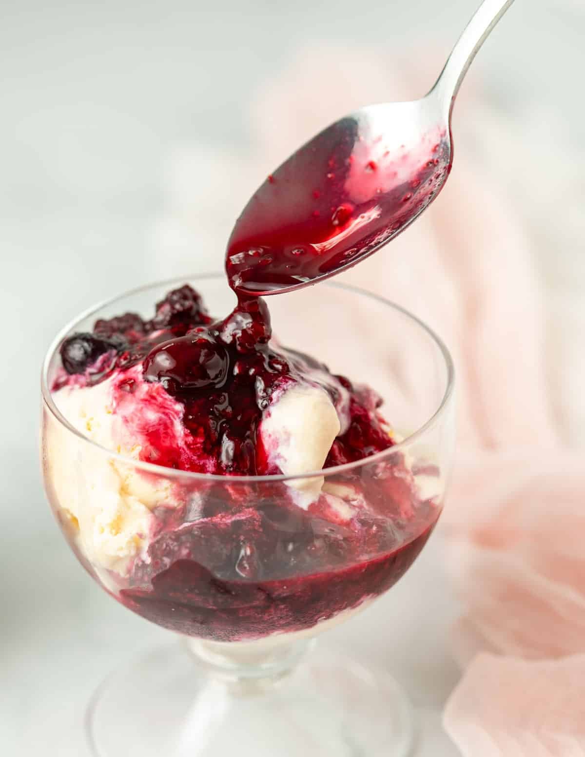 Spoon adding berry compote on top of vanilla ice cream in a bowl.
