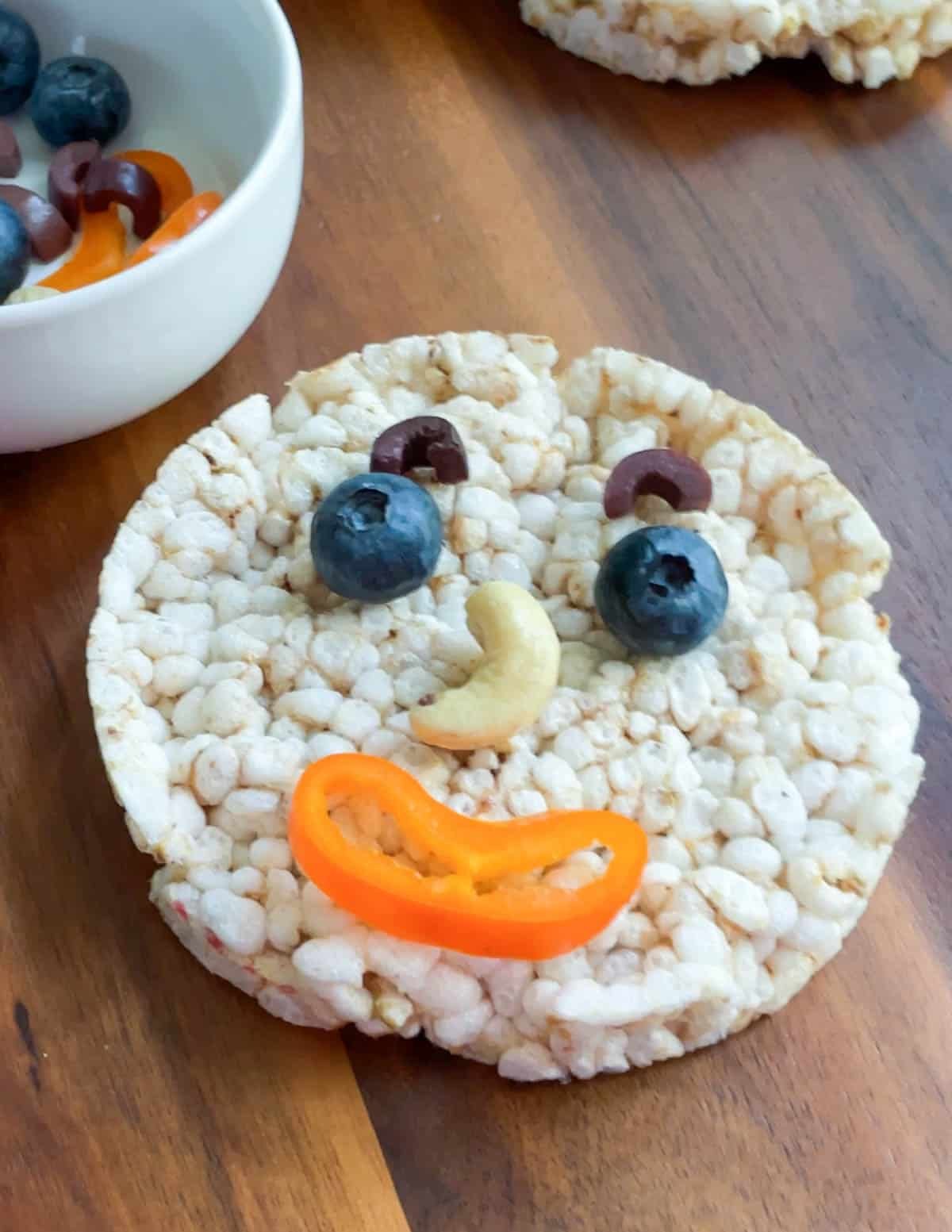 Rice cake made to look like a face with blueberry eyes, cashew nose, pepper mouth, and olive eyebrows.

