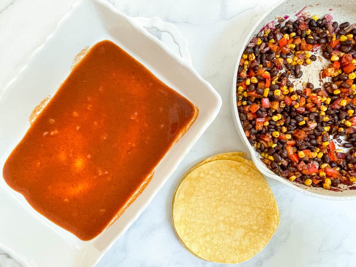 Enchilada sauce in baking dish, corn tortillas, and black bean filling.