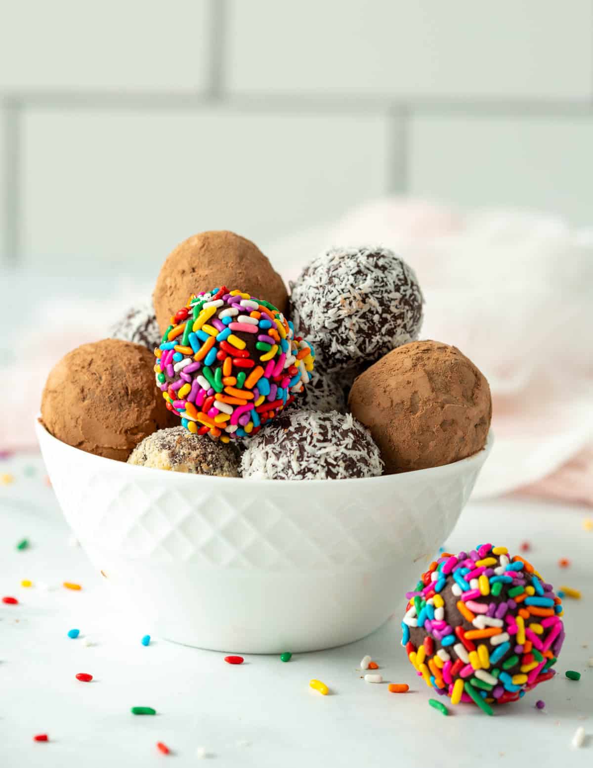 Vegan truffles in a small white bowl covered in assorted coatings: cocoa powder, coconut flakes, nuts, and sprinkles.