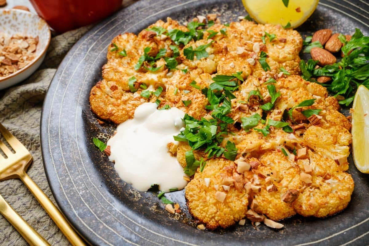 Plate of vegan cauliflower steaks served with a side of tahini and topped with almonds and parsley.
