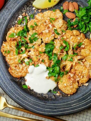 Plate of cauliflower steaks with a side of tahini and topped with parsley and nuts.