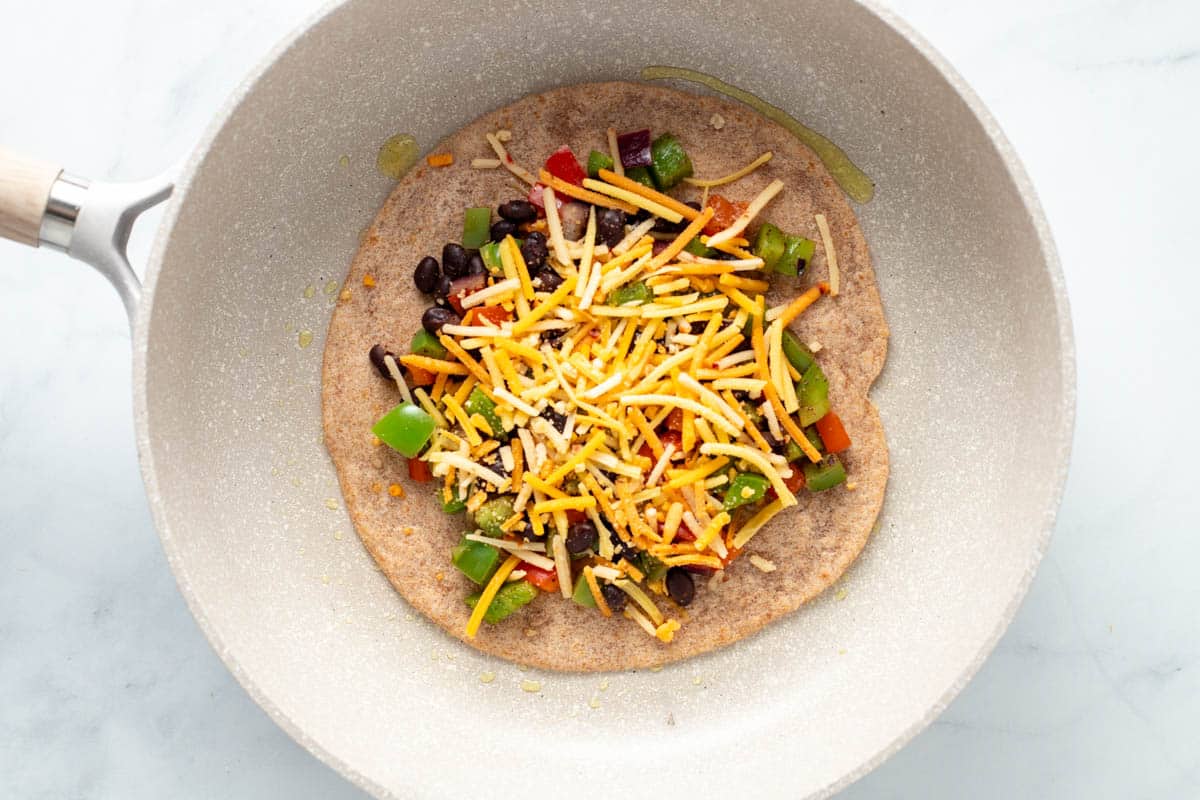 Tortilla topped with bean and vegetables and vegan cheese in a saute pan.
