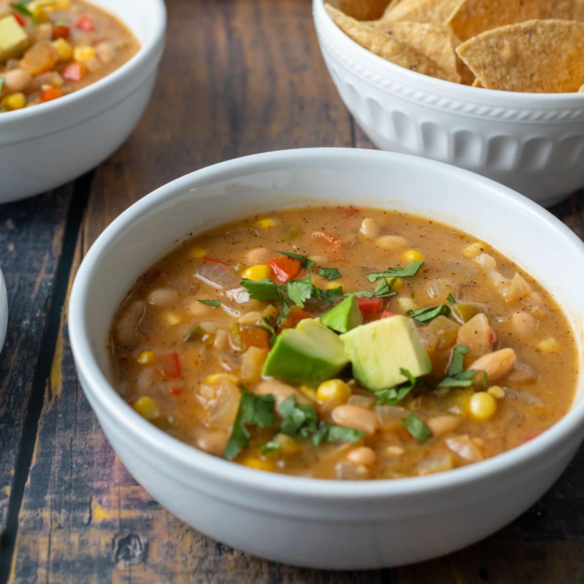 Bowl of vegan white bean chili topped with avocado and cilantro, served with a side of tortilla chips.
