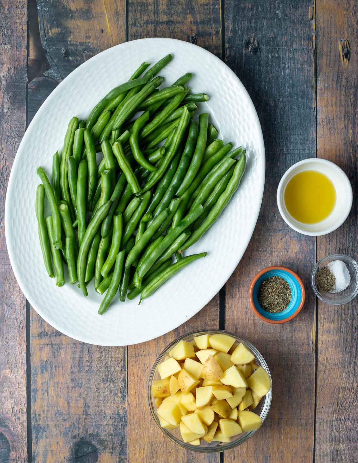 Green Beans on white plate, olive oil in small bowl, salt and pepper in small bowl, Italian seasoning in small bowl, diced Yukon gold potatoes in glass bowl.