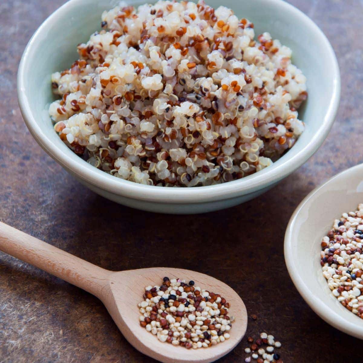 Cooked quinoa in small bowl, and uncooked quinoa on a wood spoon.