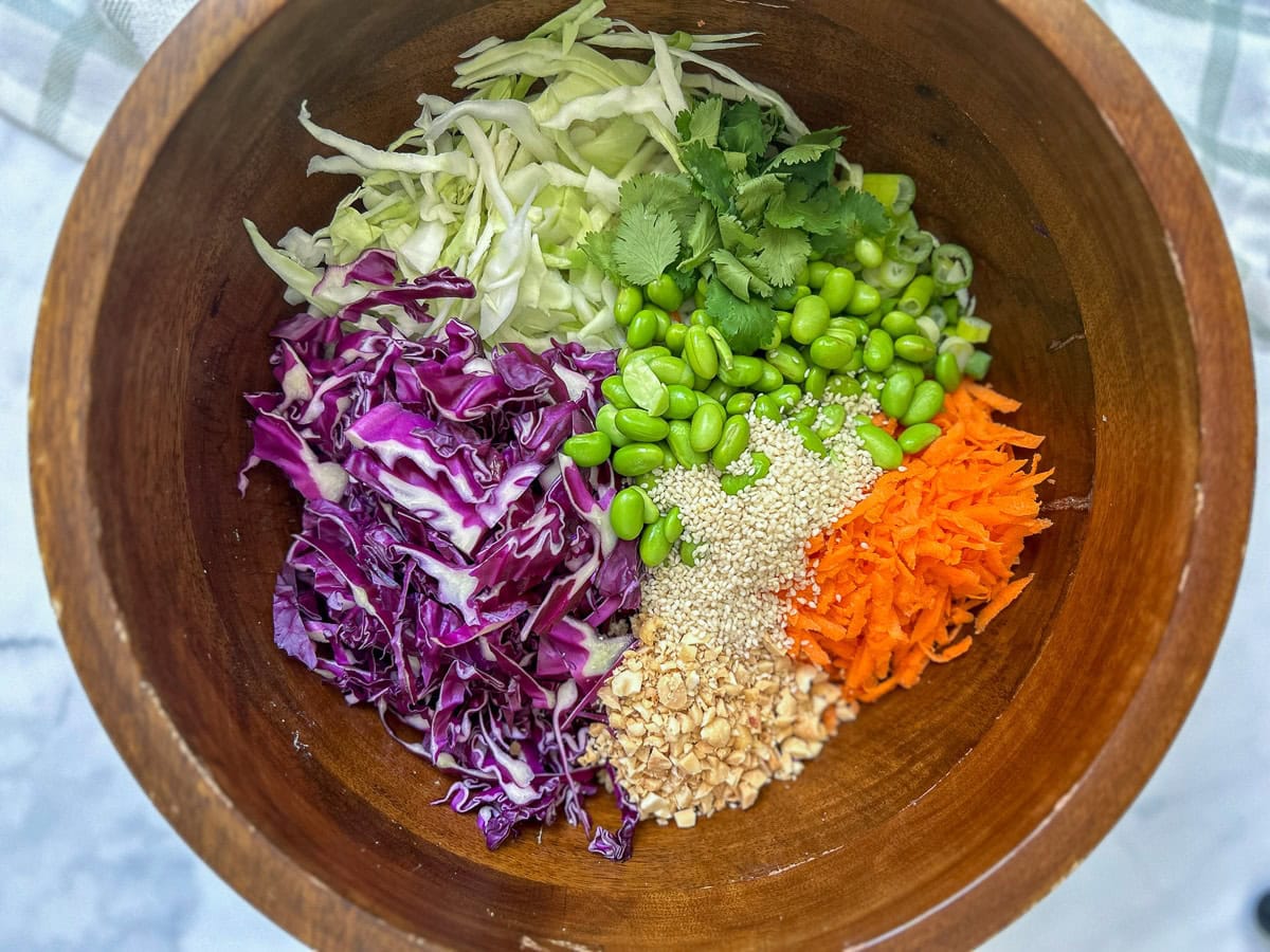 Purple cabbage, green cabbage, cilantro, green onion, carrots, peanuts, and sesame seeds in a large wood salad bowl.